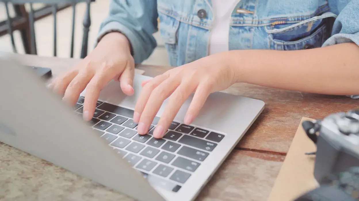 Mãos de uma pessoa a escrever num computador portátil sobre uma mesa de madeira