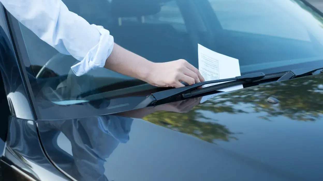 Pessoa a colocar uma notificação de multa no para-brisas de um carro preto