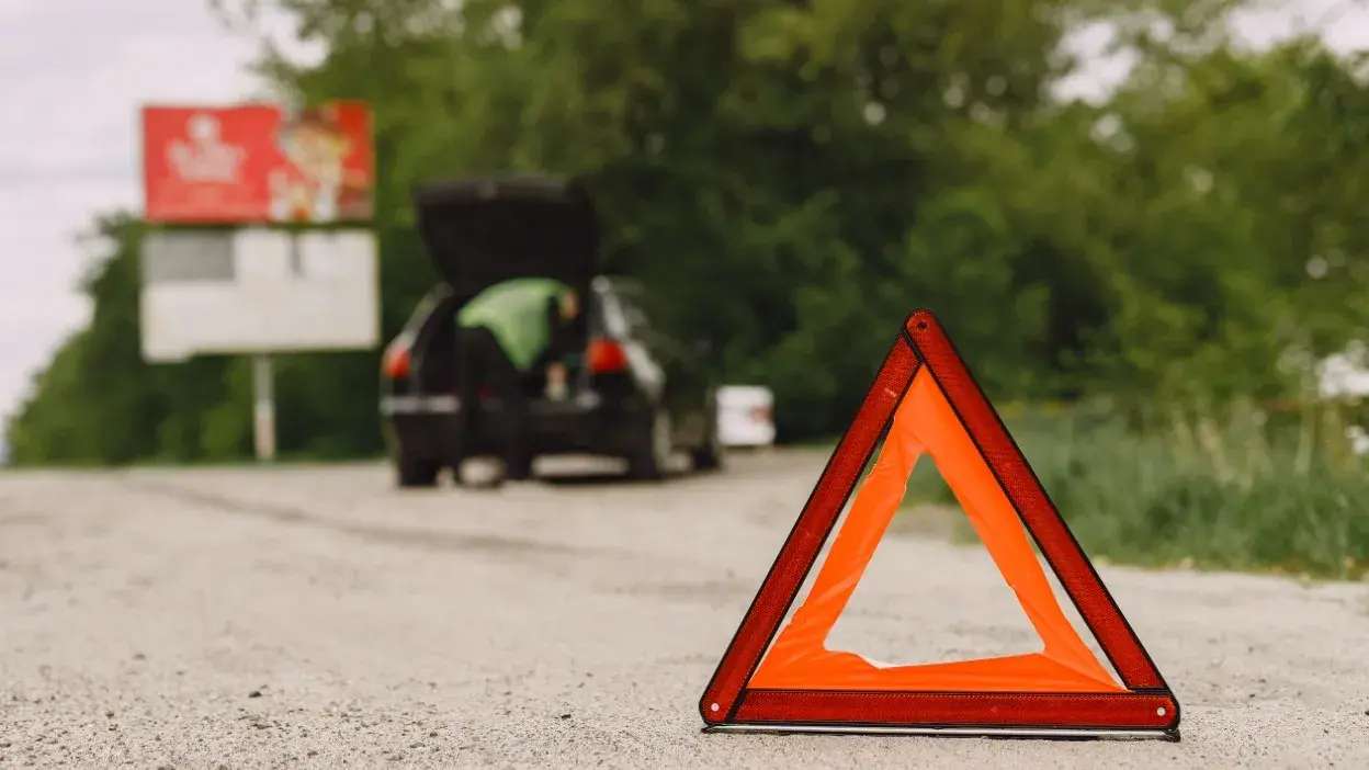 Triângulo de segurança refletor colocado na estrada com carro avariado ao fundo e pessoa a verificar o motor.